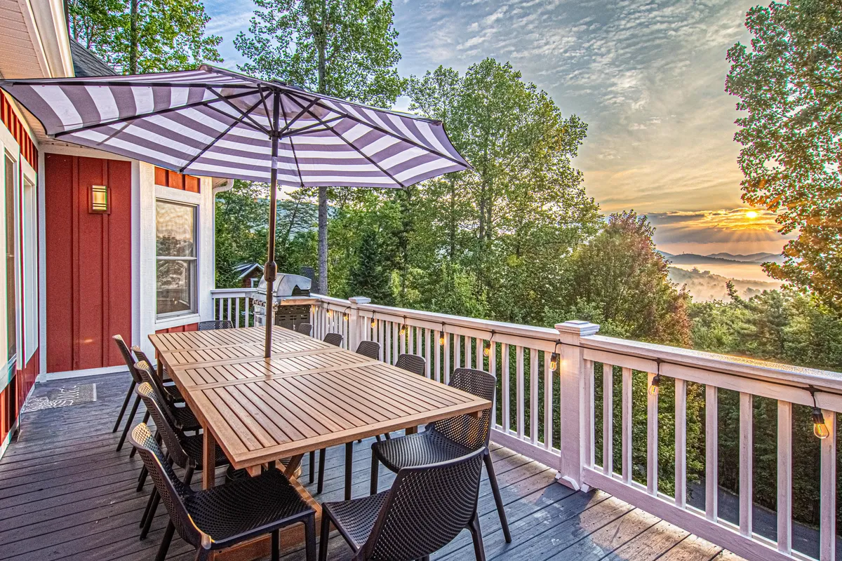 A scenic mountain-view deck outfitted with a wooden dining table beneath a striped umbrella overlooks the Blue Ridge Mountains. The inviting setup is perfect for al fresco dining, sunset watching, and gathering with friends and family in a relaxed outdoor setting.