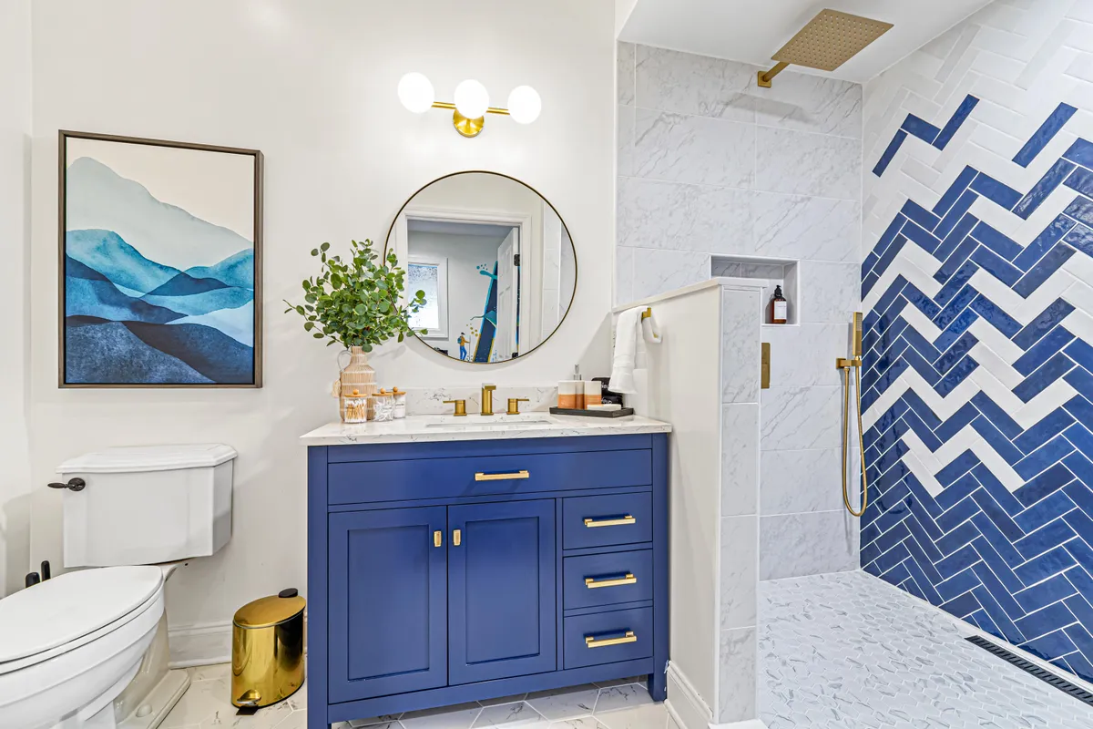 A stylish bathroom with a blue vanity, decorative mirror, and a herringbone-tile shower, all presented in a serene and inviting atmosphere. The space is well-lit and designed for personal grooming and relaxing routines, highlighting the multi-bathroom feature.