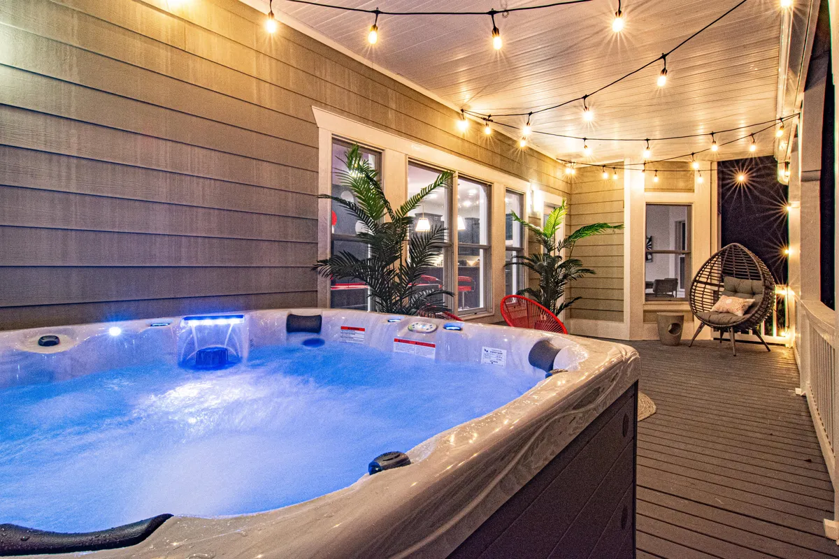 An exterior hot tub with illuminated water sits amidst lush greenery, enhanced by the ambiance of hanging string lights and a cozy hanging chair. The setting feels private and inviting, perfect for star-gazing or enjoying a warm soak under the open sky.