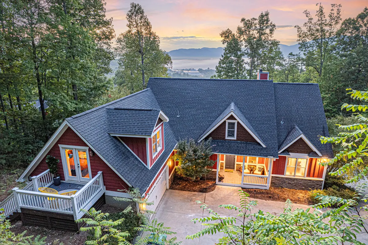 The image showcases the exterior of a spacious, luxury mountain home surrounded by lush trees. The porch, with comfortable seating, faces sweeping Blue Ridge Mountain views, and large windows hint at the inviting spaces inside. The scene radiates tranquility, perfect for savoring sunset moments or relaxing in nature.