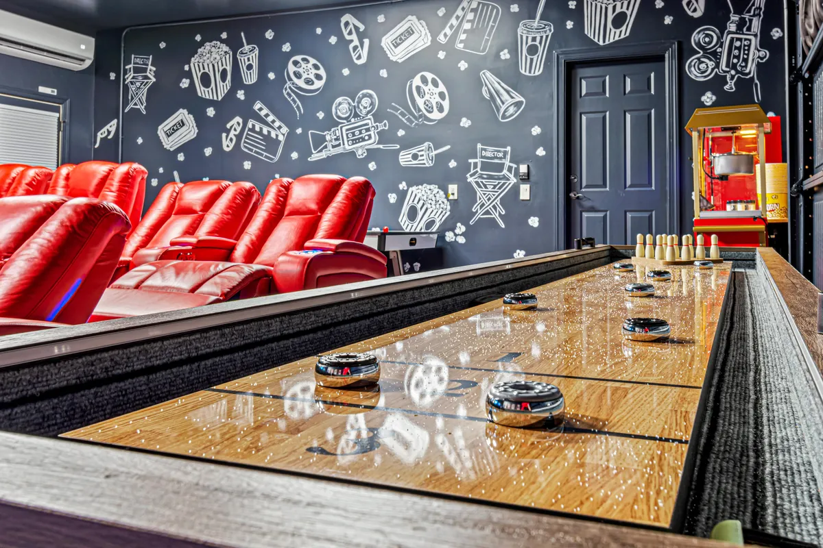 A lively entertainment area combining luxurious red reclining chairs (like those in a home theater), a shuffleboard table for friendly games, a popcorn machine, and movie-themed wall art. The overall mood is fun and relaxed, suggesting a multifunctional space for both watching movies and gaming.