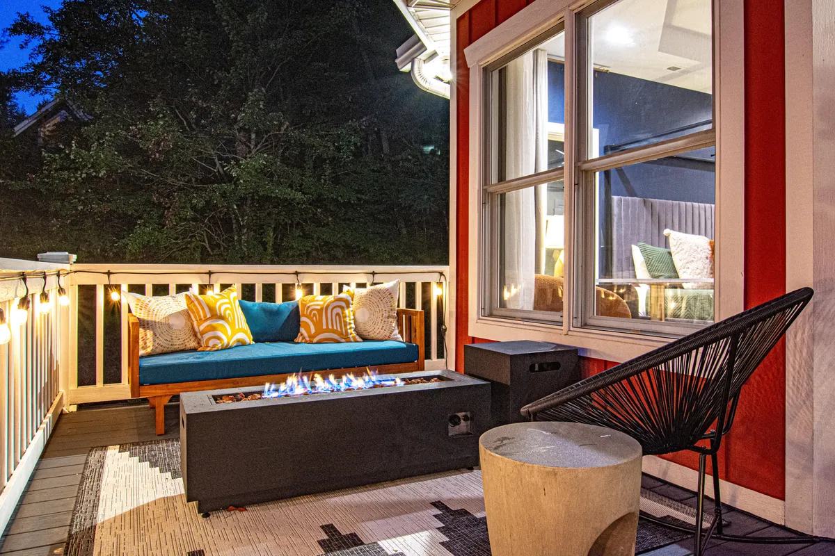 This image showcases a glowing fire pit surrounded by comfortable seating, decorative pillows, hanging lights, and a cozy sofa, all set on a spacious deck. The arrangement invites evening relaxation, social gatherings, and stargazing under a warm, welcoming ambiance.